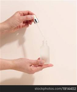 female hand holding a glass bottle and dropper for cosmetics on a beige background