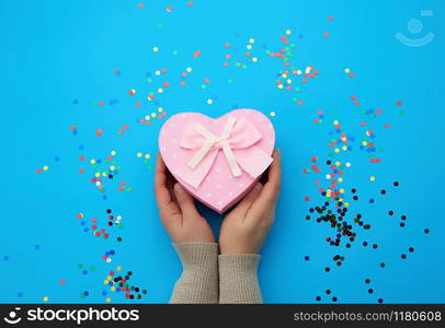 female hand holding a pink cardboard box with a bow on a blue background with multi-colored confetti, concept of giving a gift for a holiday