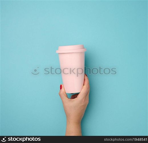 female hand holding a pink ceramic mug with a plastic lid on a blue background, break time and drink coffee
