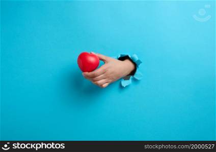 female hand holding a red heart on a blue background. Part of the body sticks out of the torn paper