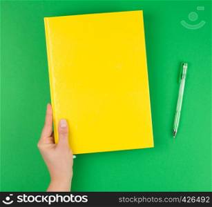 female hand holding a yellow closed notebook and pen on a green background, top view