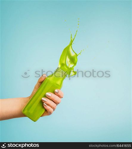 Female hand holding bottle with green splash summer beverage: smoothie or juice at blue background.