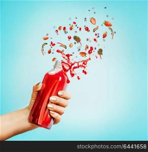 Female hand holding bottle with red splash summer beverage: smoothie or juice and berries at blue background.