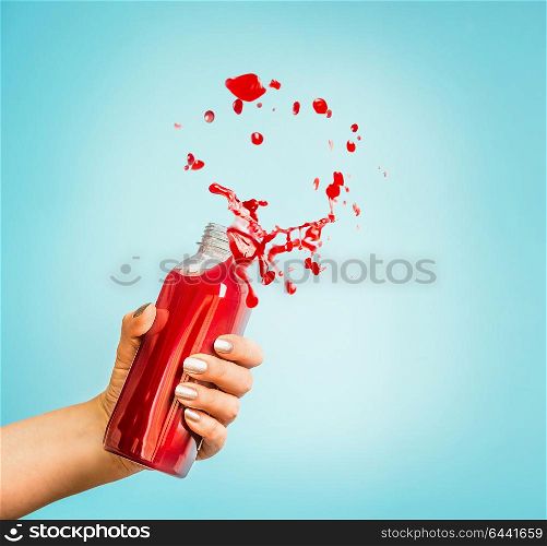 Female hand holding bottle with red splash summer beverage: smoothie or juice at blue background.