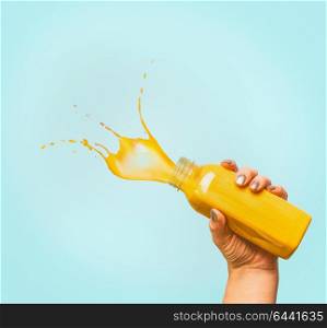 Female hand holding bottle with yellow splash summer beverage: smoothie or juice at blue background, copy space