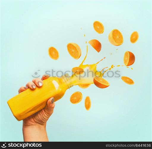 Female hand holding bottle with yellow splash summer beverage: smoothie or juice and flying oranges at blue background.