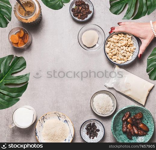 Female hand holding bowl with cashew on concrete background with vegan gluten-free cake ingredients: non dairy milk, almond flour, Coconut cream and milk, agar agar and dried fruits . Top view