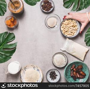 Female hand holding bowl with cashew on concrete background with vegan gluten-free cake ingredients: non dairy milk, almond flour, Coconut cream and milk, agar agar and dried fruits . Top view