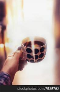 Female hand holding cup of coffee at blurred living room background with sunlight and bokeh. Copy space