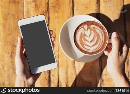 female hand holding cup of hot cocoa or chocolate while holding mobile phone on wooden table, close up