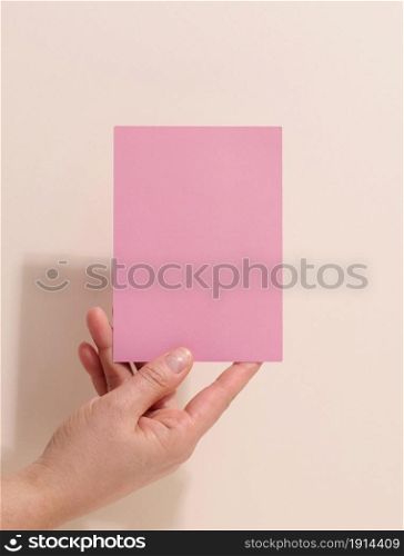 Female hand holding empty pink paper on a beige background. Copy paste image or text, close up
