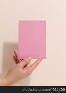 Female hand holding empty pink paper on a beige background. Copy paste image or text, close up