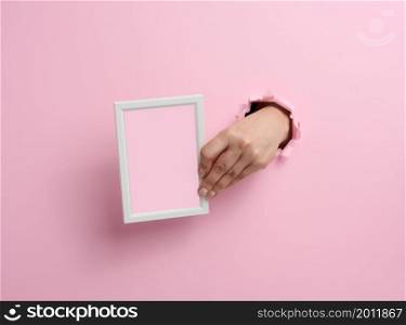 female hand holding empty white wooden frame, body part sticking out of torn hole in pink paper background