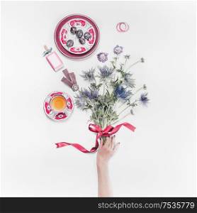 Female hand holding flowers bunch with ribbon on white desktop with cup of tea and women&rsquo;s things. Top view. Flat lay