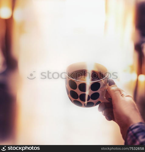 Female hand holding polka dot cup with steamed hot drink at cozy home background with sunlight and bokeh
