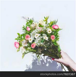 Female hand holding summer flowers bunch at sunny white background with shadow. Top view. Mothers day. Greeting bouquet