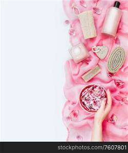Female hand holding water bowl with flowers on pink towel with various eco friendly skin care and beauty tools: brush, sponge, pumice and cosmetic products on white background. Top view. Flat lay