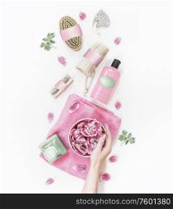 Female hand holding water bowl with pink flowers on white desktop with various eco friendly spa and skin care tools and cosmetic product bottles. Top view. Modern natural cosmetic. Zero waste
