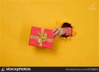female hand holds a box with a gift on a yellow background, part of the body sticks out of a torn hole in a paper background. Congratulation, holiday surprise