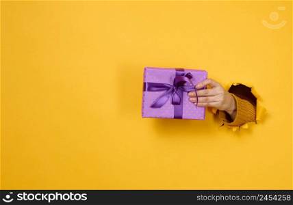 female hand holds a box with a gift on a yellow background, part of the body sticks out of a torn hole in a paper background. Congratulation, holiday surprise 