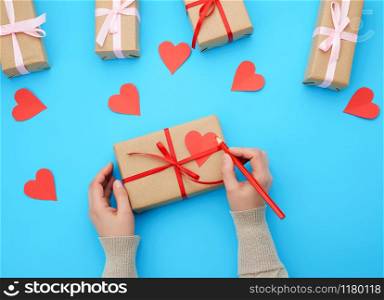 female hand holds a box wrapped in brown kraft paper and tied with a silk ribbon on a blue background, paper red hearts. Valentine&rsquo;s Day backdrop
