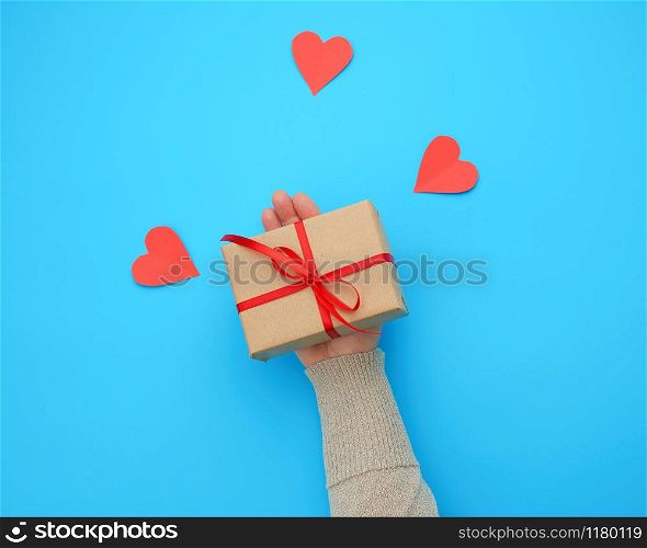 female hand holds a box wrapped in brown kraft paper and tied with a silk ribbon on a blue background, paper red hearts. Valentine&rsquo;s Day backdrop