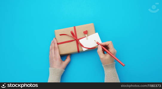 female hand holds a box wrapped in brown kraft paper and tied with a silk red ribbon on a blue background, paper red hearts. Valentine&rsquo;s Day backdrop