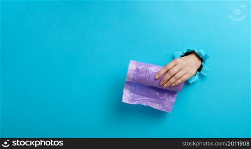 female hand holds a bundle of transparent garbage bags on a blue background. A part of the body sticks out of a torn hole in the paper, copy space