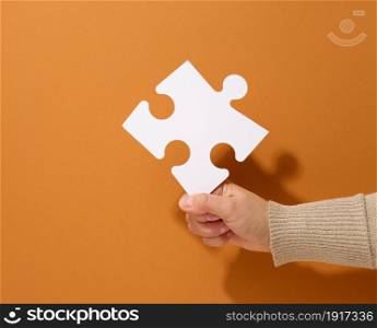 female hand holds a fragment of a white large puzzle on a brown background, the concept of finding an idea, solving a problem