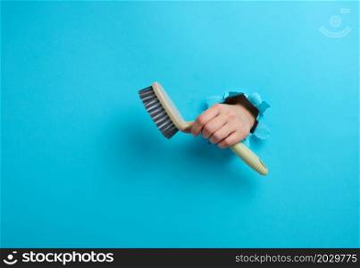 female hand holds a gray plastic brush on a blue background. A part of the body sticks out of a hole with torn edges in a paper background