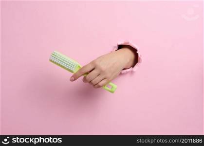 female hand holds a green plastic brush on a pink background. A part of the body sticks out of a hole with torn edges in a paper background