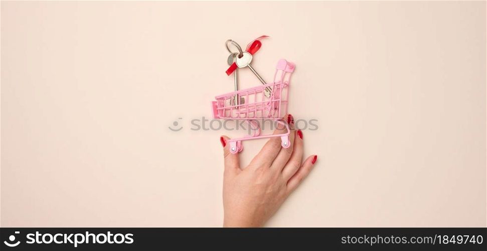 female hand holds a miniature metal shopping cart with keys on a beige background. Real estate purchase, mortgage, discount, banner