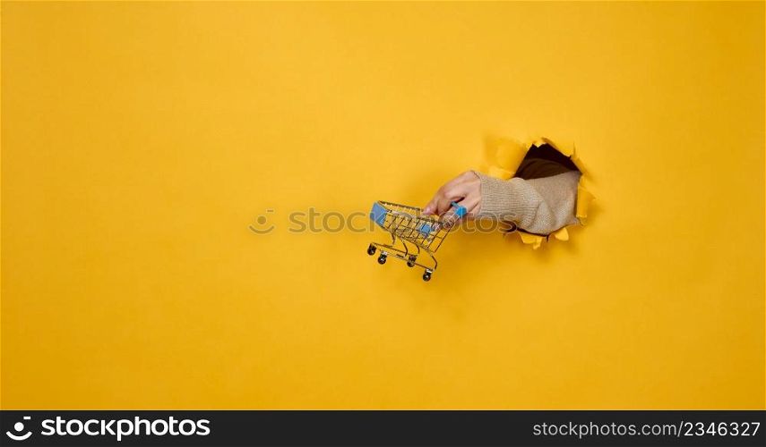 female hand holds a miniature shopping cart on a yellow background, a part of the body sticks out of a torn hole in a paper background. Business concept, start of sales and online shopping