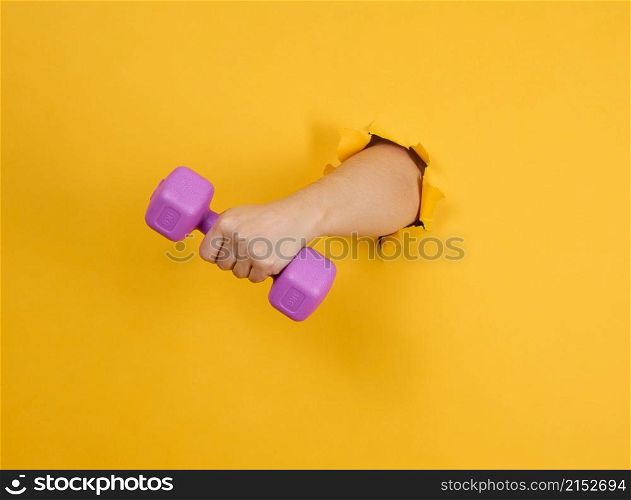 female hand holds a plastic kilogram dumbbell on a yellow background, a part of the body sticks out of a torn hole in a paper background. Healthy lifestyle, sports