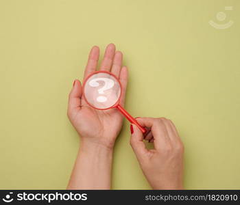 female hand holds a plastic magnifying glass and question marks on a green background. The concept of finding an answer to questions, truth and uncertainty.