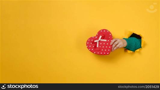 female hand holds a red box with a gift on a yellow background, part of the body sticks out of a torn hole in a paper background. Congratulation, holiday surprise. Copy space