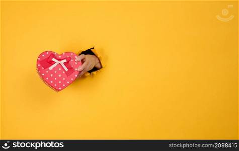 female hand holds a red box with a gift on a yellow background, part of the body sticks out of a torn hole in a paper background. Congratulation, holiday surprise. Copy space