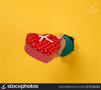 female hand holds a red box with a gift on a yellow background, part of the body sticks out of a torn hole in a paper background. Congratulation, holiday surprise