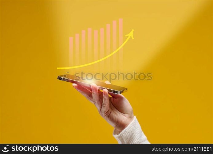 female hand holds a smartphone with holographic graphics on a yellow background. Performance growth, success