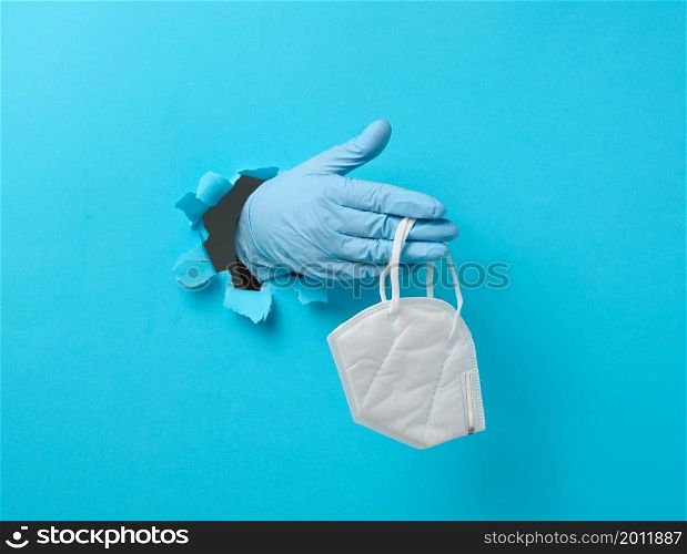 female hand holds a white disposable medical mask to protect against viruses during an epidemic and pandemic. Part of the body sticks out of a torn hole in blue paper