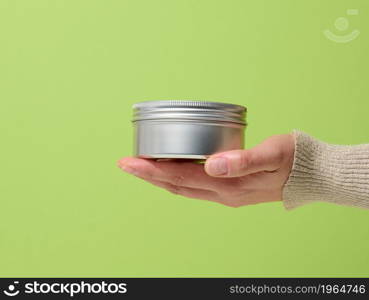 female hand holds an aluminum jar for cream on a green background. Container for cosmetics