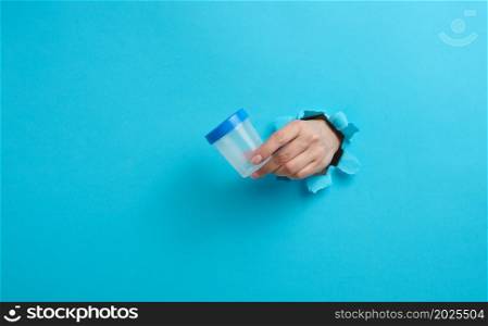 female hand holds an empty plastic jar for urine tests, the hand is sticking out of a torn hole in a blue background