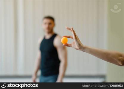 Female hand holds ping pong ball, man in sportswear on background, table-tennis training game in gym. Table tennis concept