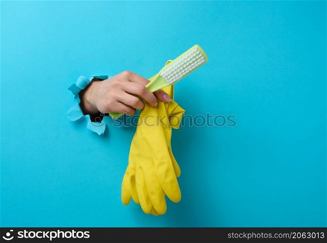 female hand holds rubber gloves and a plastic cleaning brush, part of the body sticks out of a torn hole in a blue paper background