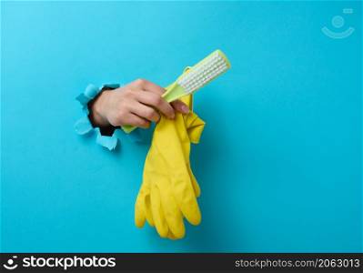 female hand holds rubber gloves and a plastic cleaning brush, part of the body sticks out of a torn hole in a blue paper background