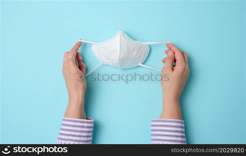 female hand holds white disposable medical face mask against viral diseases, blue background, top view