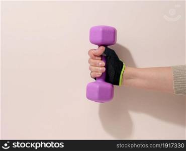 female hand in a black sports glove holds a purple one kilogram dumbbell on a beige background