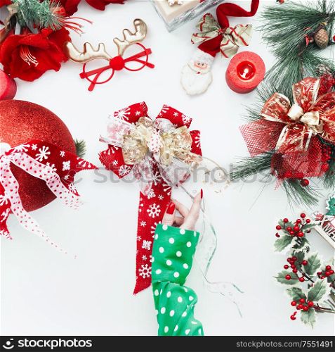 Female hand in green blouse holding Christmas bow on white desktop with red holiday decoration. Top view