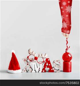 Female hand in red sweater with white snowflakes holding ribbon of Christmas ball. Christmas background with Santa hut, word Xmas, candles and red holiday ball stand on light gray background. Modern