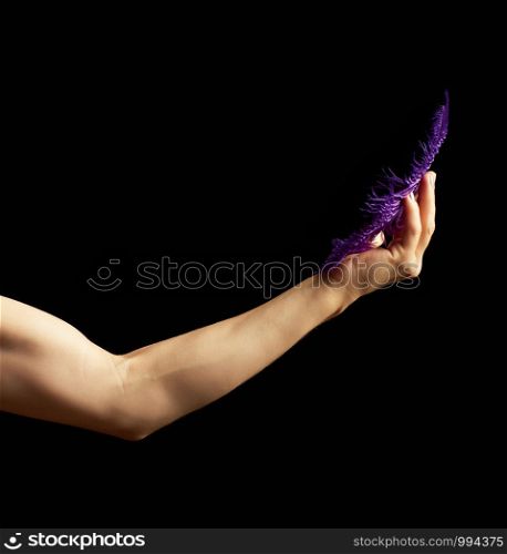 female hand is bent at the elbow, the muscles are tense and hold a blue feather on a dark background, concept of light and heavy